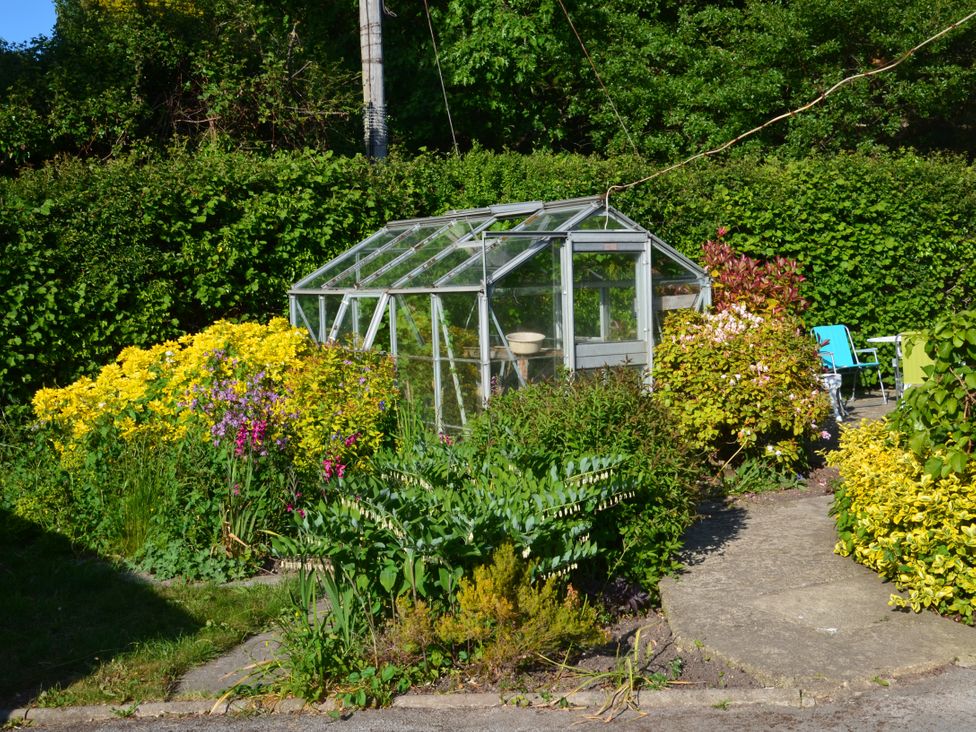 A garden with a small glass greenhouse surrounded by various plants and flowers with a seating area in the background