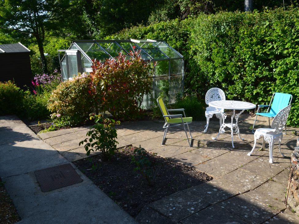 A patio area with a white metal table and chairs and a green and blue garden chairs near a greenhouse surrounded by plants at Woodstock in Chaffcombe near Chard