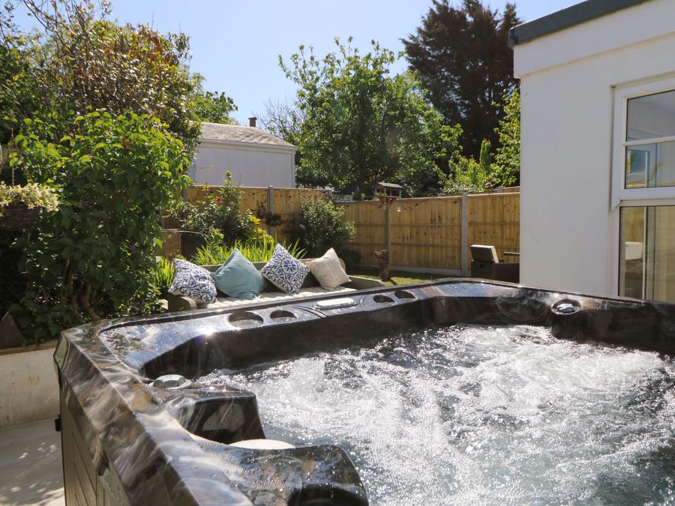 A bubbling hot tub next to a white building with a garden couch and pillows in a fenced backyard at Towyn Hall in New Quay