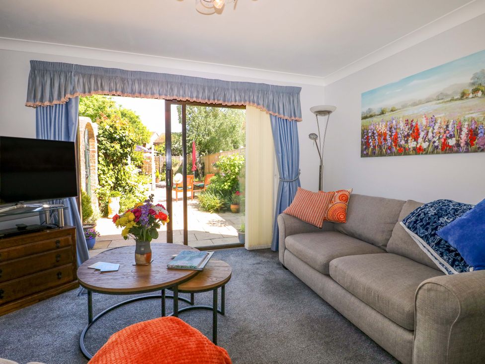 A living room with a gray sofa pillows a round wooden coffee table with flowers a television on a wooden dresser and sliding glass doors leading to a garden at Lavender Cottage in Hailsham