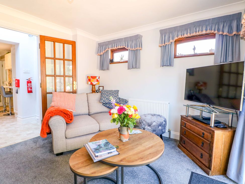 A living room with a sofa and pillows coffee tables with flowers a television on a wooden stand and curtains at Lavender Cottage in Hailsham