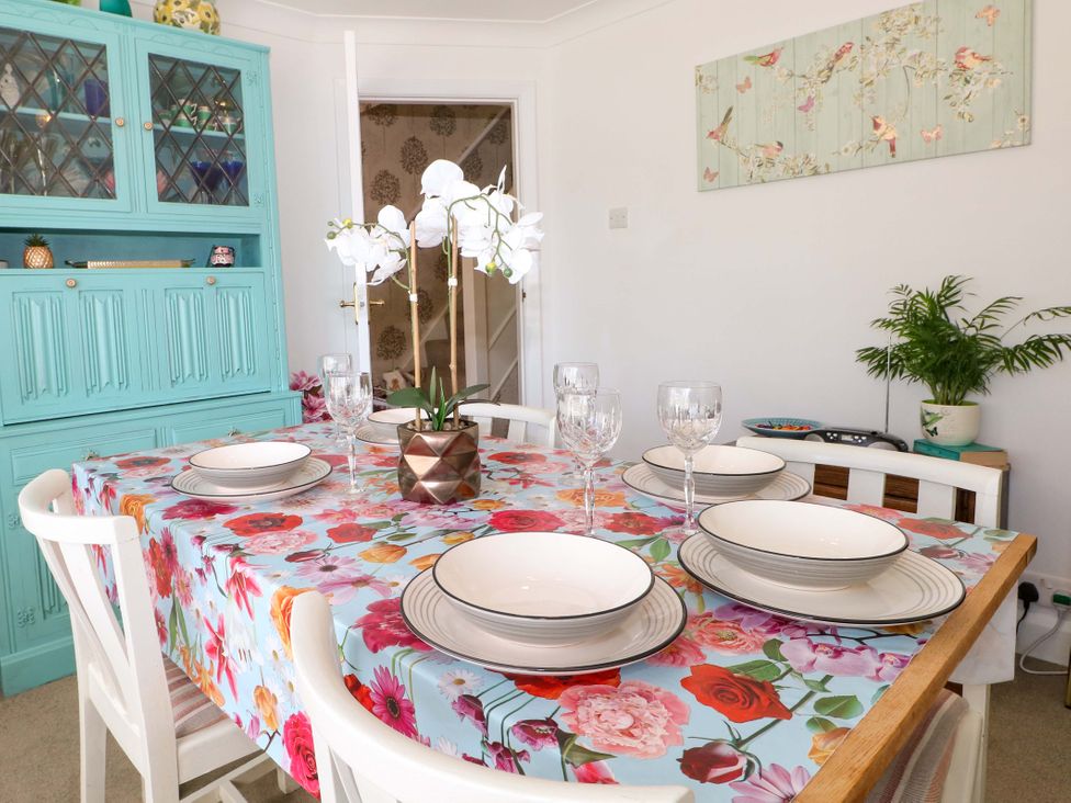 A dining room with a floral tablecloth on a wooden table set with plates glasses and a white orchid in a pot with a turquoise cabinet and a potted plant on a small table at Lavender Cottage in Hailsham