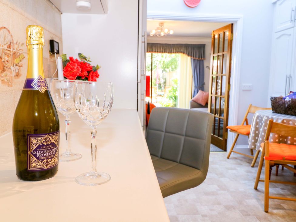 A kitchen counter with a bottle of prosecco and two wine glasses next to a grey chair and dining area at Lavender Cottage in Hailsham