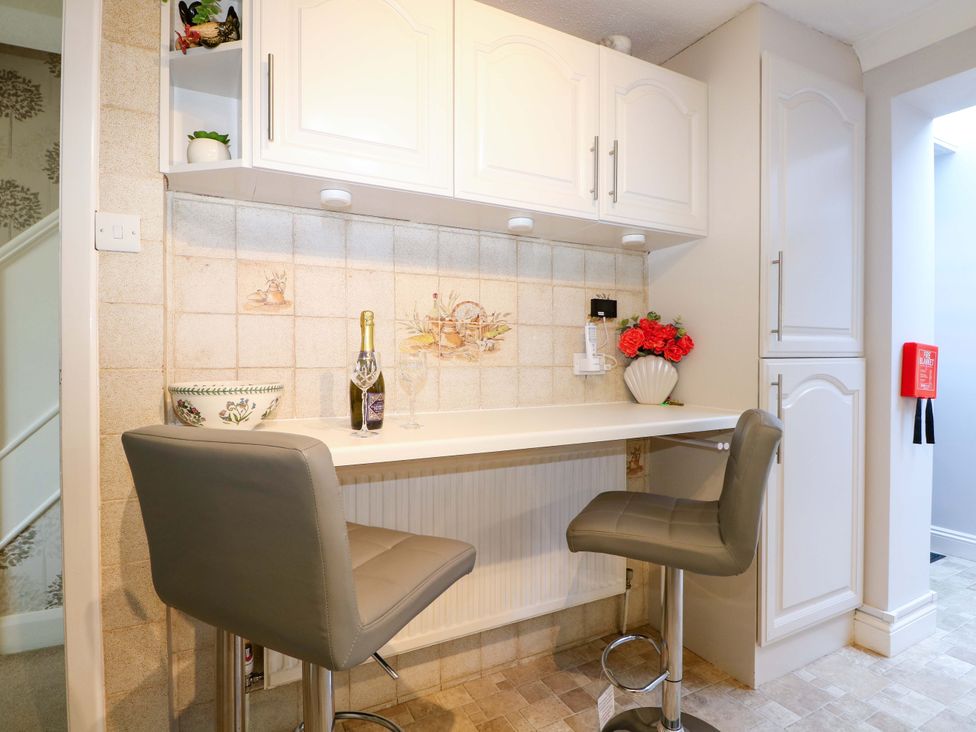 A kitchen corner with bar counter two grey bar stools a bottle with two glasses flowers in a vase and white cabinets at Lavender Cottage in Hailsham