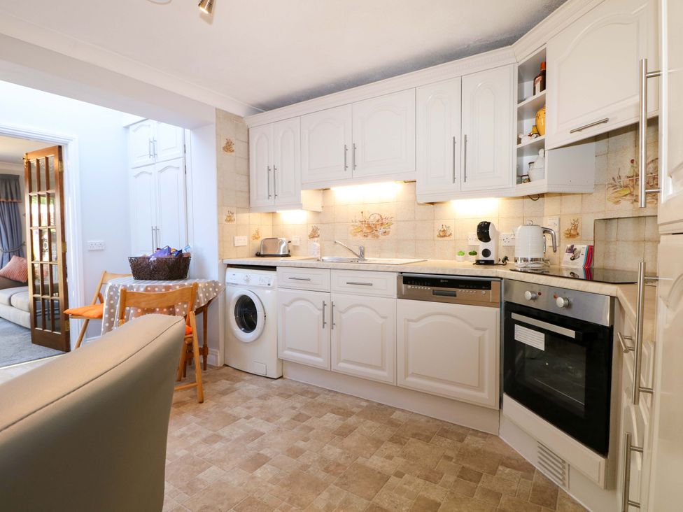 A kitchen with white cabinets a washing machine an oven a small table with chairs and a view into a living room at Lavender Cottage in Hailsham