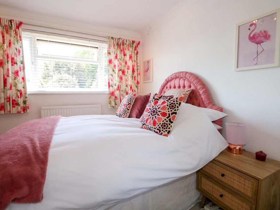 A bedroom with a bed with a pink headboard pillows floral curtains and a wooden nightstand with a lamp at Lavender Cottage in Hailsham