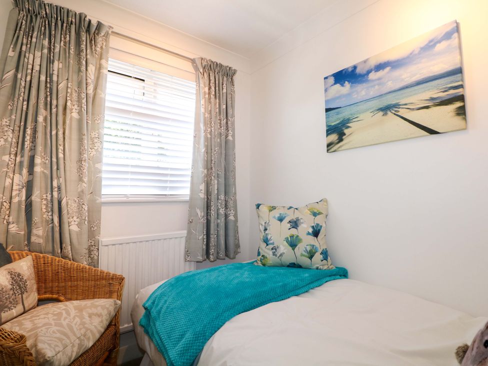 A bedroom with a single bed with a floral pillow and blue blanket a wicker chair a window with patterned curtains and a beach photo on the wall at Lavender Cottage in Hailsham
