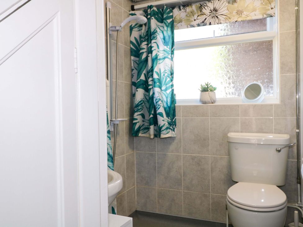 A bathroom with a toilet a sink a shower with a tropical shower curtain and a window with a plant and mirror on the sill at Lavender Cottage in Hailsham