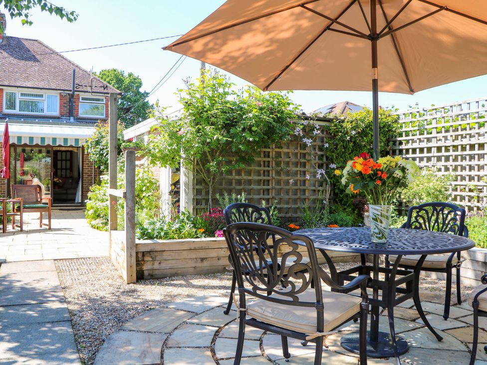 An outdoor garden area with a round metal table and chairs under a large umbrella with flowers in a vase on the table at Lavender Cottage in Hailsham