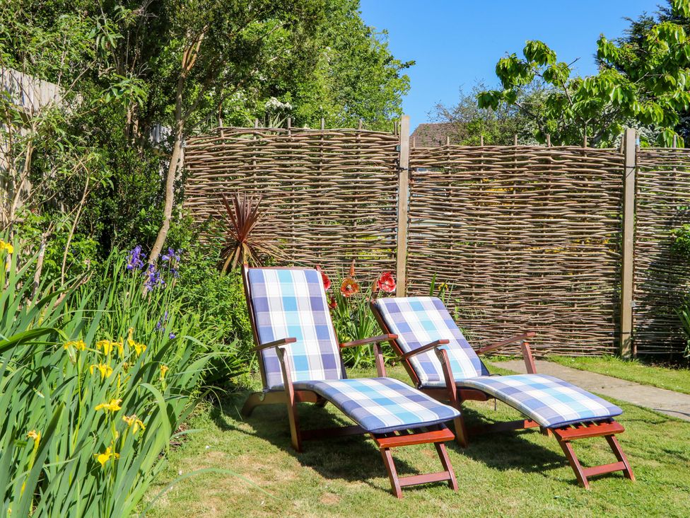 Two wooden lounge chairs with blue and white plaid cushions on a lawn by a woven fence and garden at Lavender Cottage in Hailsham