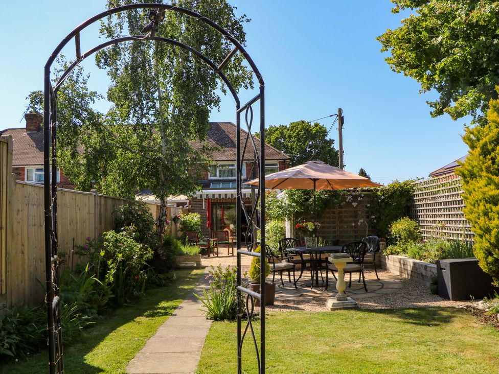 A garden with a metal archway, a paved path, a patio with a table and chairs under an umbrella, plants and shrubs at Lavender Cottage in Hailsham