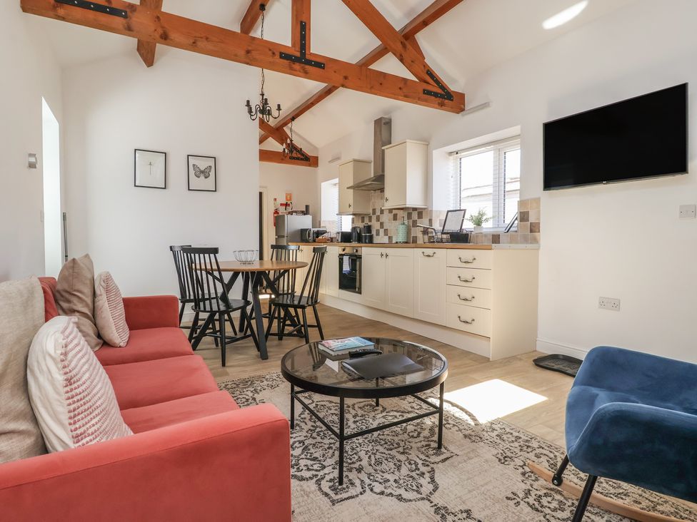 A living area with a red sofa, round glass coffee table, dining table with black chairs, kitchen cabinets, and a wall-mounted TV at Guillemot in Flamborough