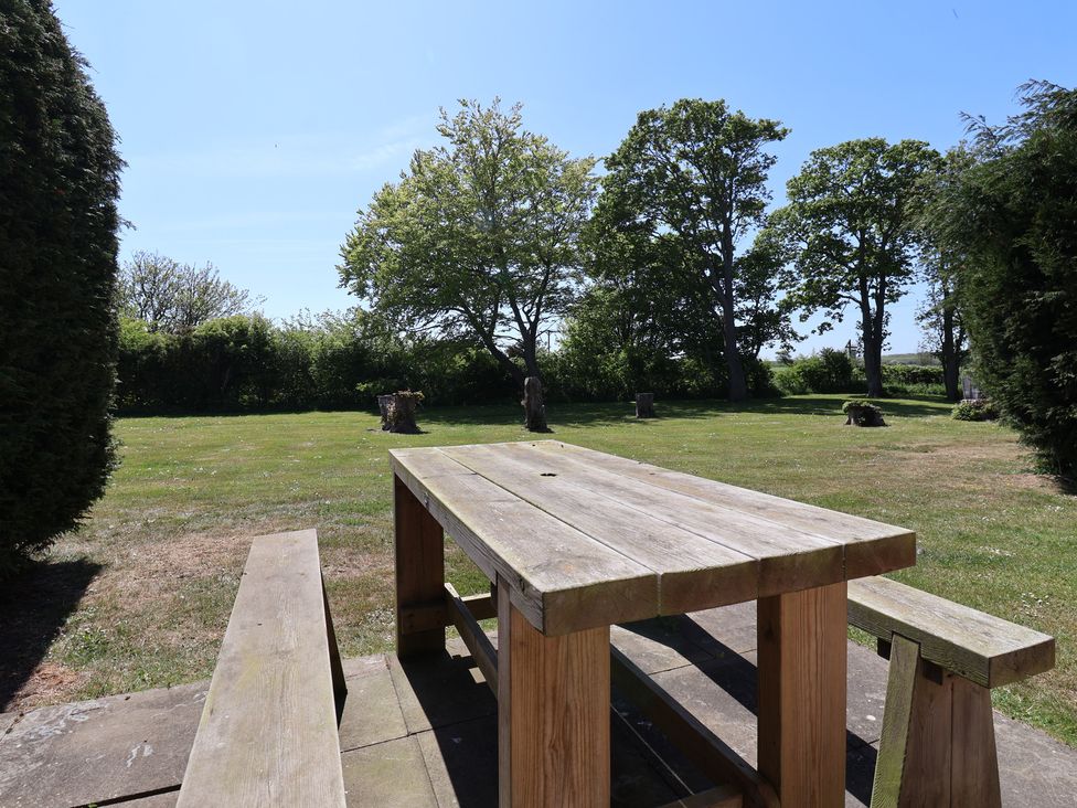 An outdoor wooden picnic table with benches on a paved area surrounded by grass and trees at Guillemot in Flamborough