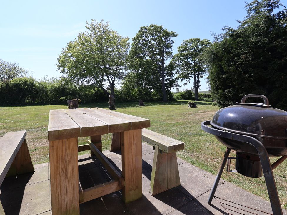 A wooden picnic table with benches on a patio and a charcoal grill in a grassy garden area with trees in the background at Guillemot in Flamborough