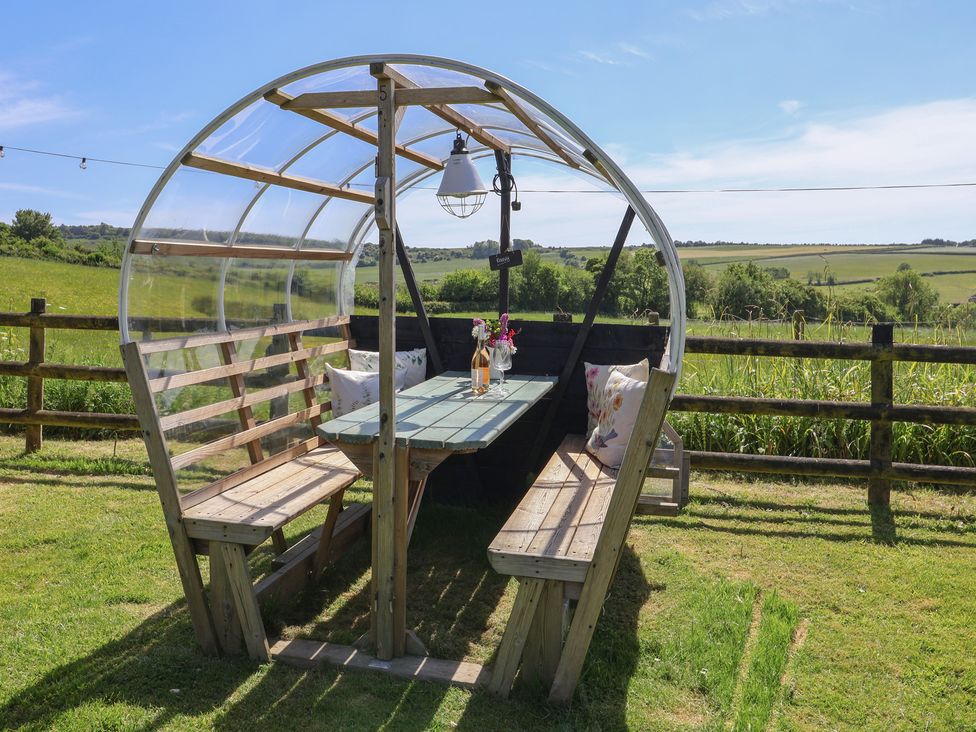 An outdoor wooden seating area with a table and cushions under a transparent canopy in a grassy field at Eggardon Rest in Askerswell near Bridport