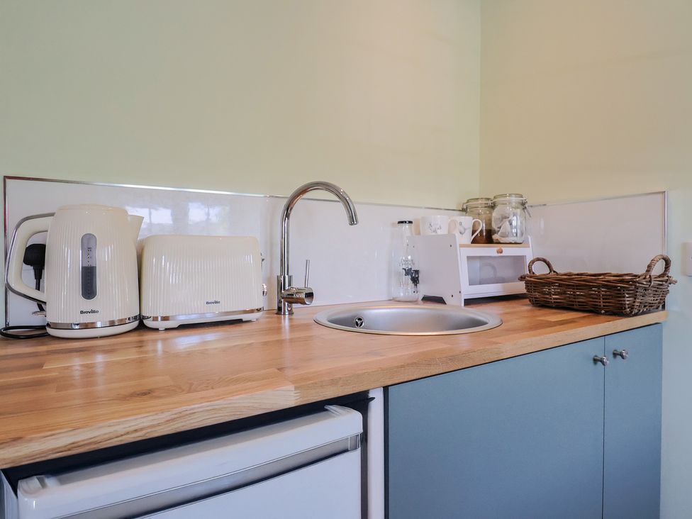 A kitchen counter with a kettle, toaster, sink, glass bottle, microwave, jars, mugs, and a wicker basket at Eggardon Rest in Askerswell near Bridport