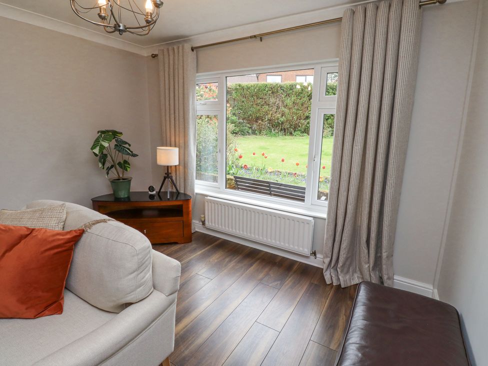 A living room with a beige sofa red cushion wooden corner table with a plant and lamp large window with beige curtains and wooden floor at Sparrow House in Alnwick