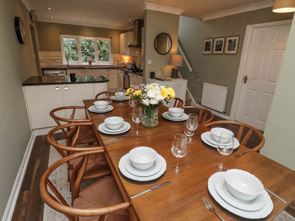 A dining area with a wooden table set with white dishes and glassware and a kitchen with counters and a window at Sparrow House in Alnwick