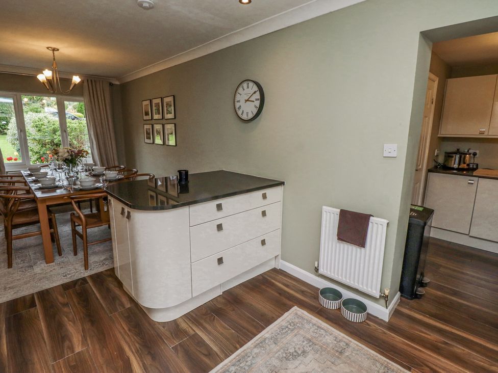 A dining area with a wooden table set with dishes and glasses next to a kitchen island with drawers and a wall clock at Sparrow House in Alnwick
