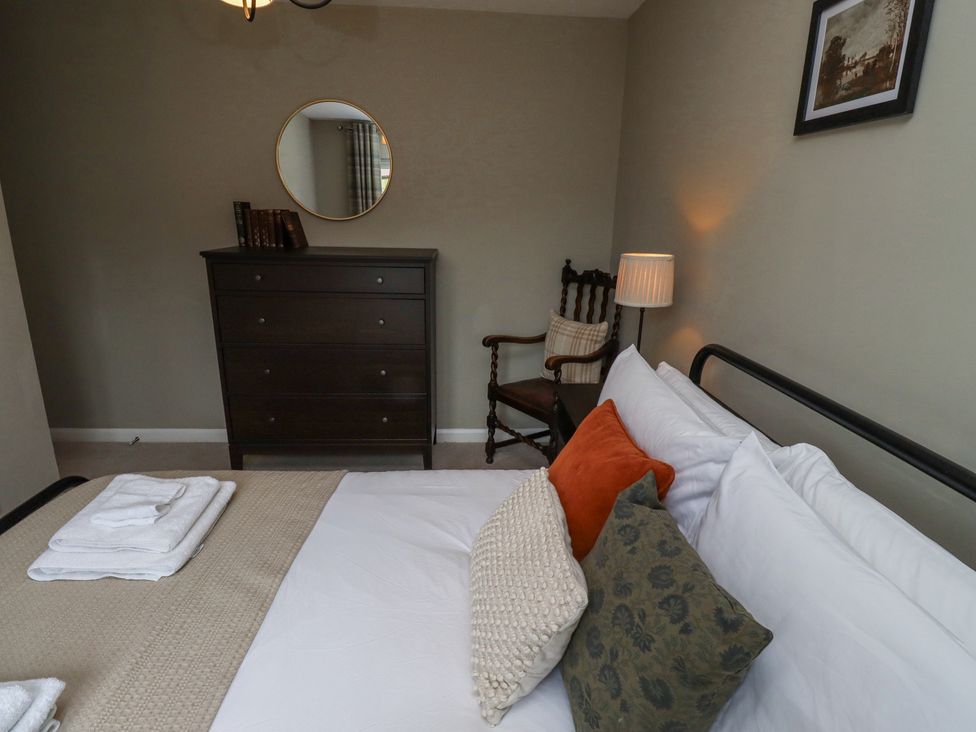 A bedroom with a bed featuring multiple pillows and folded towels a wooden dresser with books and a round mirror above a wooden chair with a cushion a bedside lamp and a framed picture on the wall at Sparrow House in Alnwick