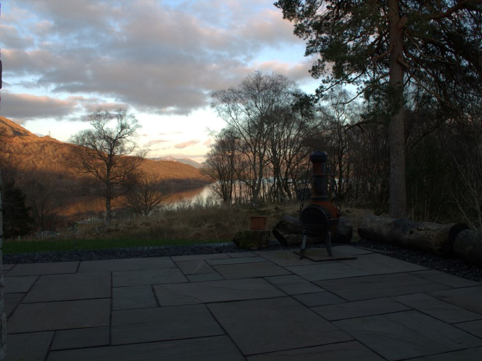 A patio with a stove and logs beside a lake at Taigh Creag Thulach in Taynuilt