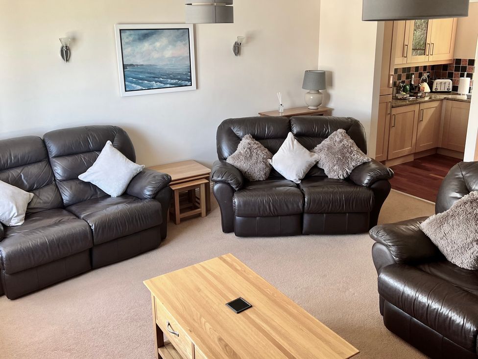 A living room with three brown leather sofas with cushions a wooden coffee table and side tables and a kitchen visible through an open doorway at Penpraze in Portreath