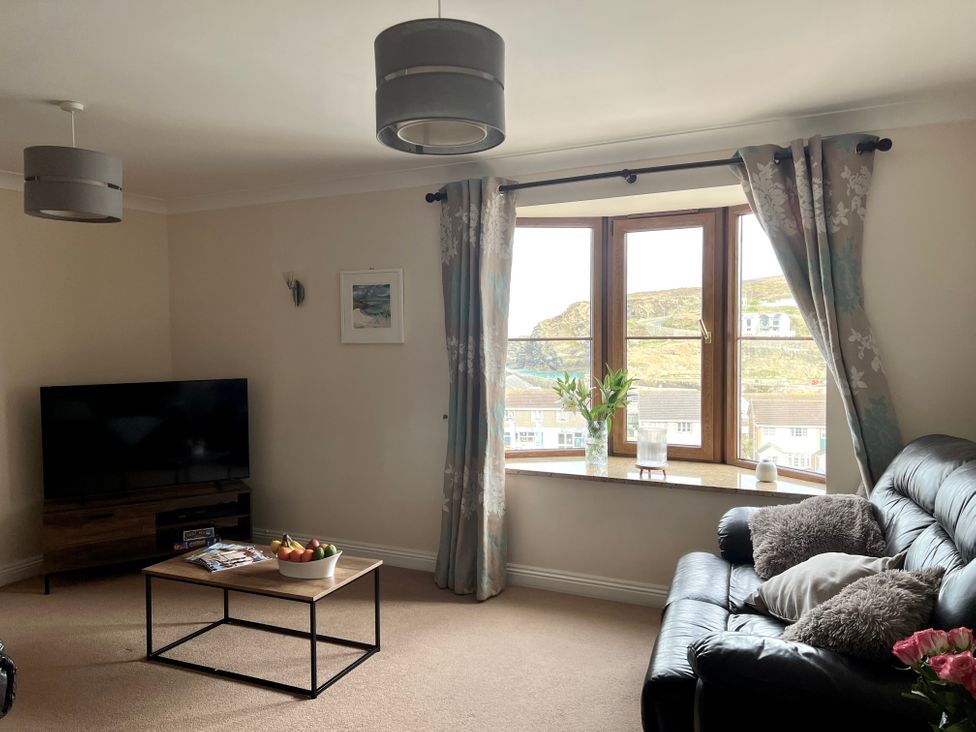A living room with a black leather sofa a wooden coffee table with a bowl of fruit a television on a stand and a window with curtains at Penpraze in Portreath