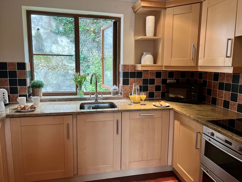 A kitchen with wooden cabinets a window above the sink croissants on a plate mugs a toaster a microwave a plate with cookies and glasses of orange juice at Penpraze in Portreath