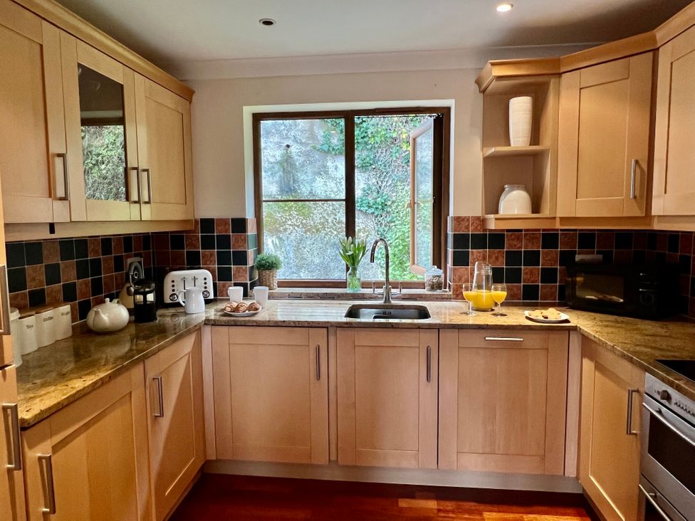 A kitchen with wooden cabinets a sink under a window and a countertop with a toaster a coffee maker cups and a jug with orange juice at Penpraze in Portreath