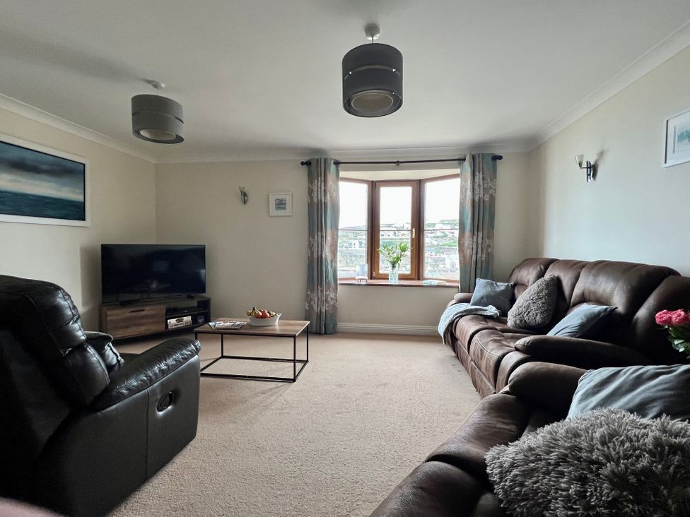 A living room with leather sofas a coffee table a television on a stand and a bay window with curtains and flowers at Curnows in Portreath