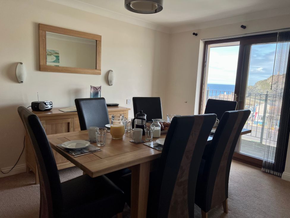 A dining room with a wooden table set for breakfast and black chairs near a glass door with a sea view at Curnows in Portreath