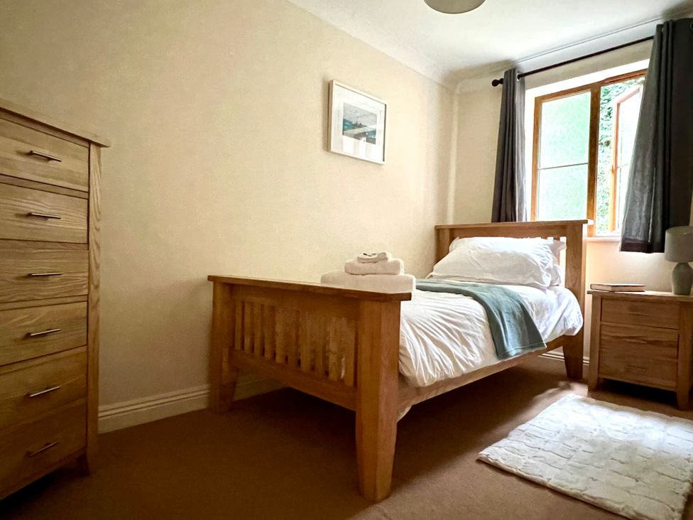 A bedroom with a single wooden bed with white bedding and blue throw a wooden chest of drawers a wooden bedside table with a lamp and window with grey curtains at Curnows in Portreath