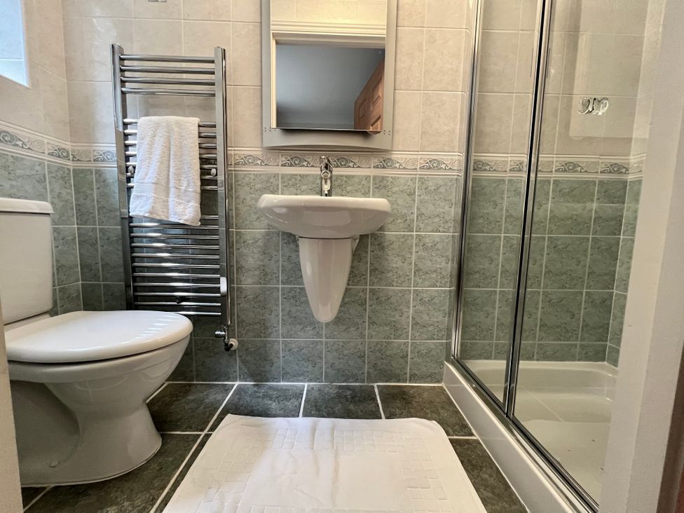 A bathroom with a toilet, a sink with a mirror above, a towel rack with a towel, a shower with glass doors, and a white bath mat on the floor at Curnows in Portreath
