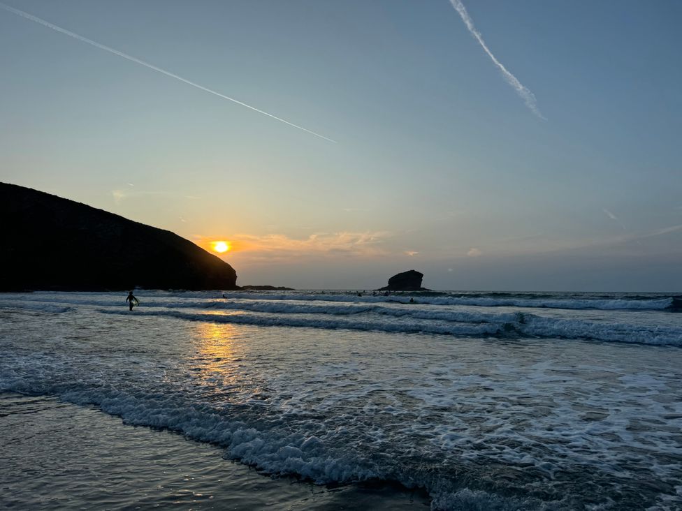 A beach at sunset with waves and a person carrying a surfboard in the water at Curnows Portreath