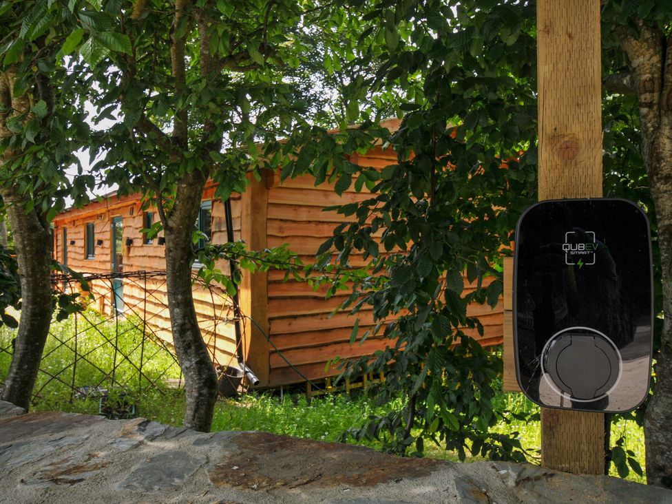 An electric vehicle charger with a wooden building behind at Owl Lodge in Menheniot