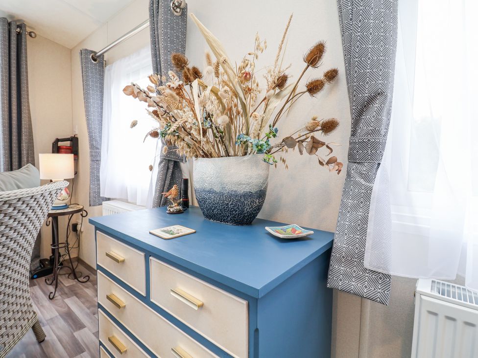 A chest of drawers with a blue top and beige drawers with gold handles and a vase with dried flowers on it next to two windows with gray curtains at The Hoot in Great Finborough near Stowmarket