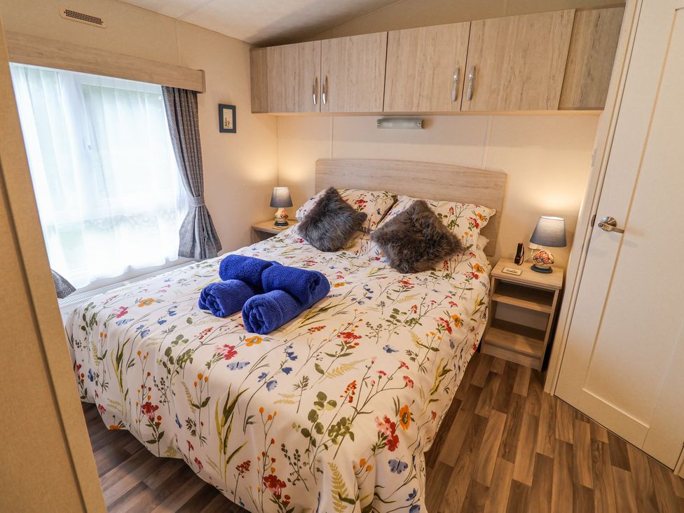A bedroom with a bed with floral bedding blue towels on the bed two bedside tables with lamps and overhead cabinets at The Hoot in Great Finborough near Stowmarket