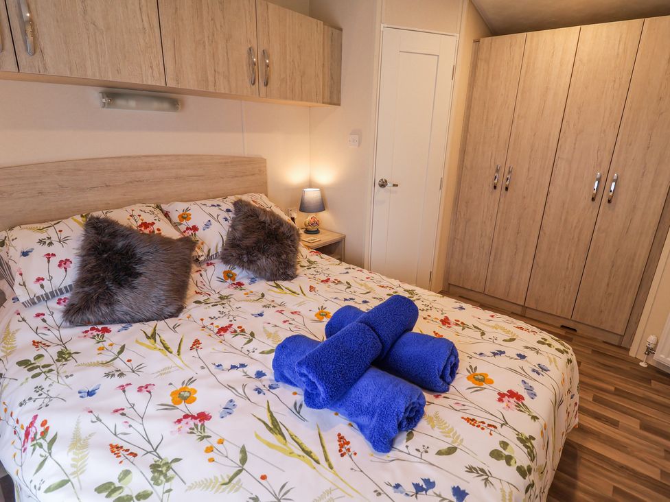 A bedroom with a floral bedspread blue towels on a bed fur pillows wooden cabinets and a bedside table lamp at The Hoot in Great Finborough near Stowmarket