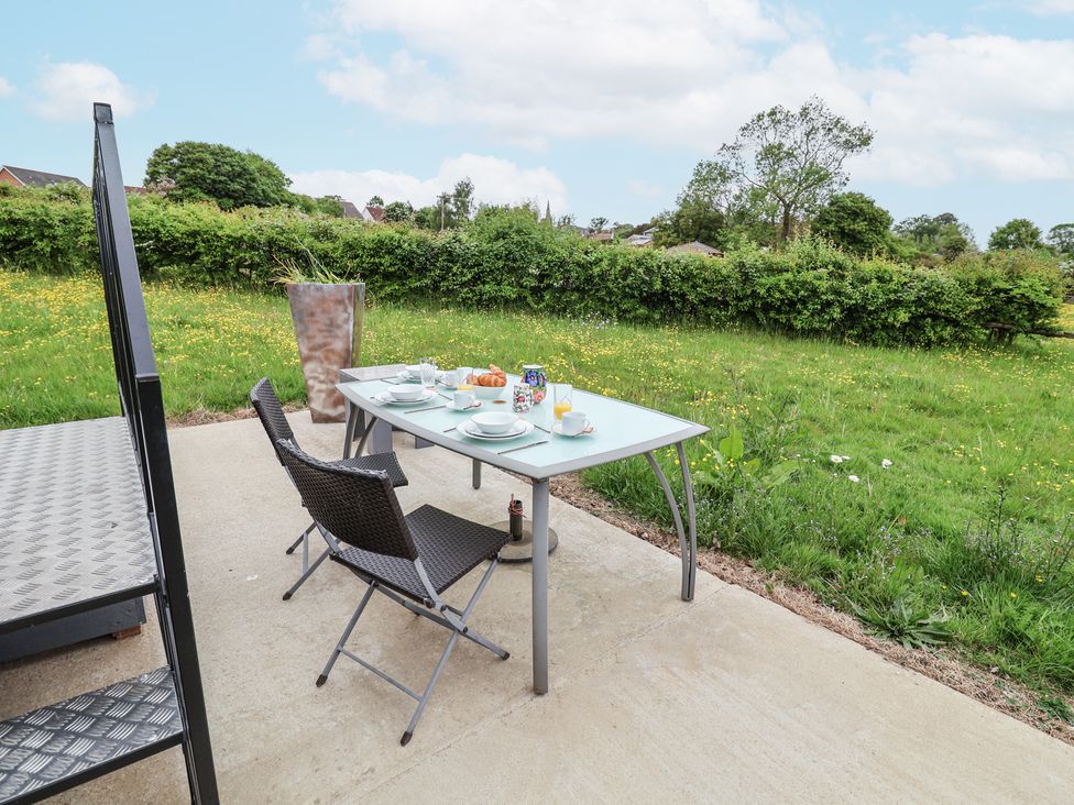 An outdoor dining table with two chairs on a patio next to a grassy field with trees at The Hoot in Great Finborough near Stowmarket
