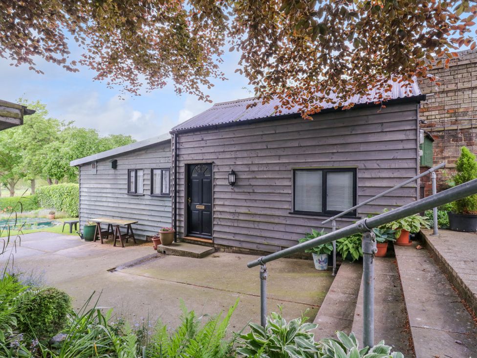 An exterior view of a small wooden building with a black door and window and potted plants on a concrete patio at The Cottage in Sudbury