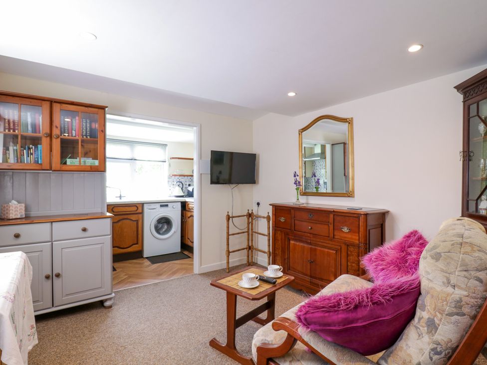 A living room with a floral armchair and purple cushions a wooden cabinet with a mirror and a bookshelf visible next to a kitchen at The Cottage in Sudbury