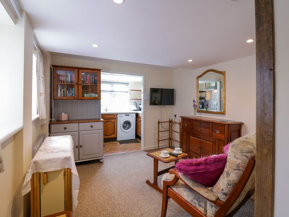A sitting area with a chair and table with cups next to a wooden sideboard with mirror and a TV on the wall at The Cottage in Sudbury