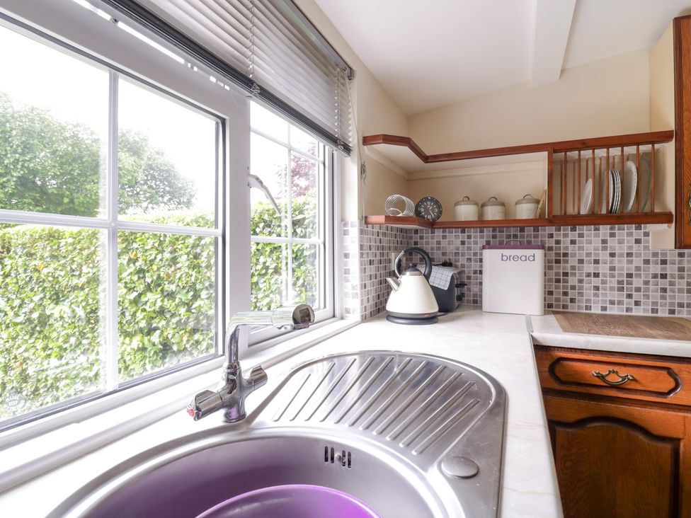 A kitchen sink with a faucet next to a window and a countertop with a kettle bread box and shelf with jars and plates at The Cottage in Sudbury