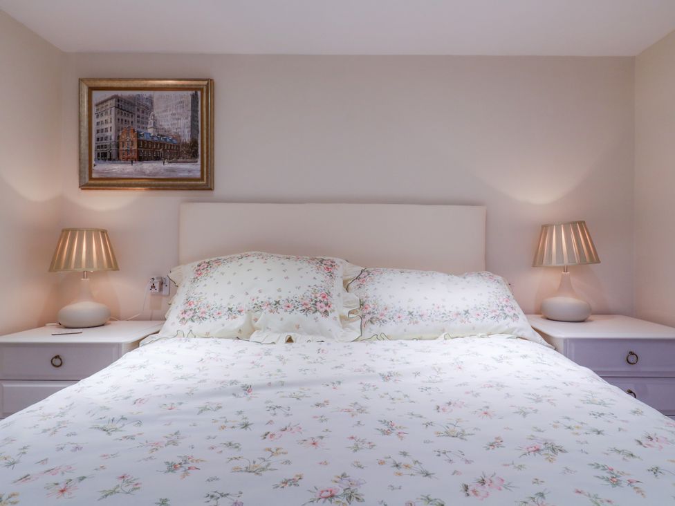 A bedroom with a bed with floral bedding and two bedside tables with lamps and a framed picture on the wall at The Cottage in Sudbury