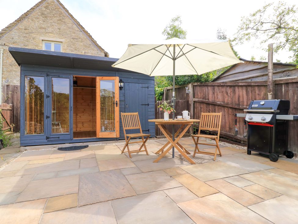 A patio with a table and chairs under an umbrella a barbecue and a wooden shed at Bramley Cottage near Cirencester