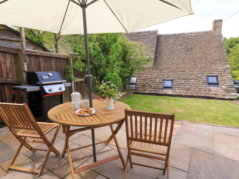 A patio with wooden table and chairs under umbrella next to barbecue grill and garden at Bramley Cottage near Cirencester
