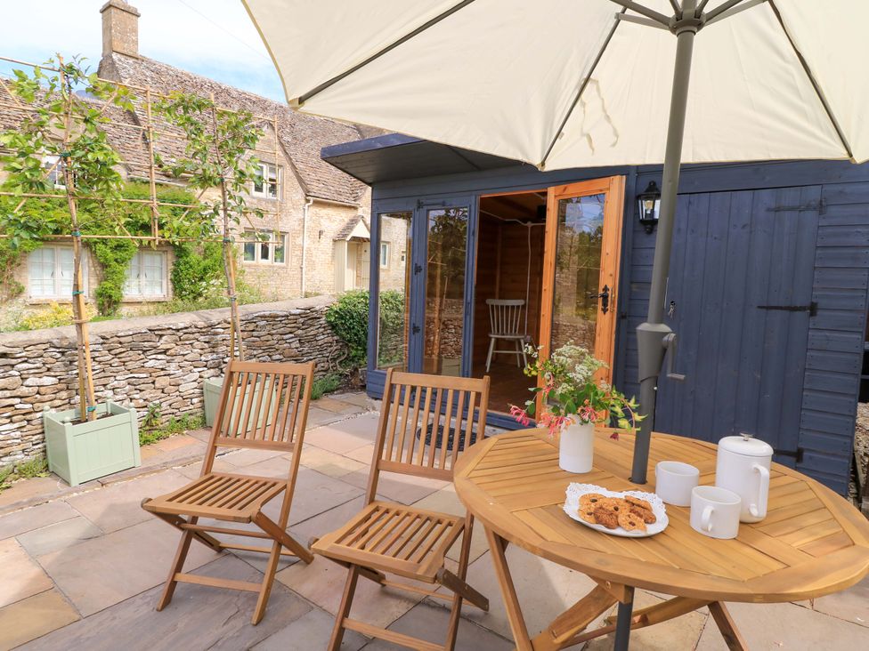 An outdoor patio with wooden chairs and table under an umbrella and a dark blue wooden shed near Cirencester