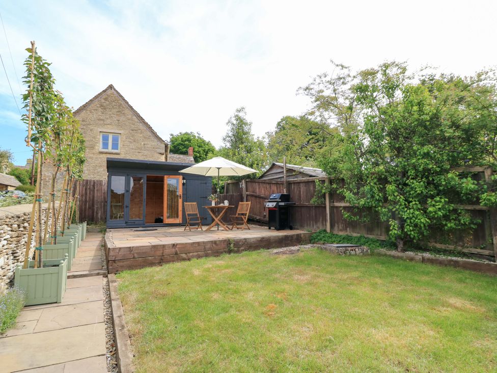 A garden with a wooden patio table and chairs under an umbrella a barbecue and a small wooden outbuilding at Bramley Cottage near Cirencester