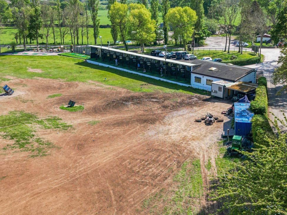An outdoor driving range with covered golf bays a parking area and open field with equipment at The Buck in Llanerch Park near St Asaph