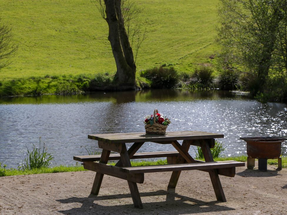 A wooden picnic table with a flower basket on top next to a body of water with grass and trees in the background at Hollie in Raglan